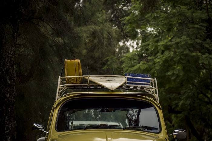 Photograph of a yellow vintage car with a roof rack. The roof rack contains a suitcase and a surfboard.