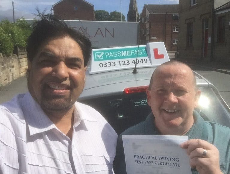 A selfie of a PassMeFast driving instructor and student driver. They are stood in front of a car with a PassMeFast advertising roof box. The learner driver is holding a practical driving test pass certificate and smiling.