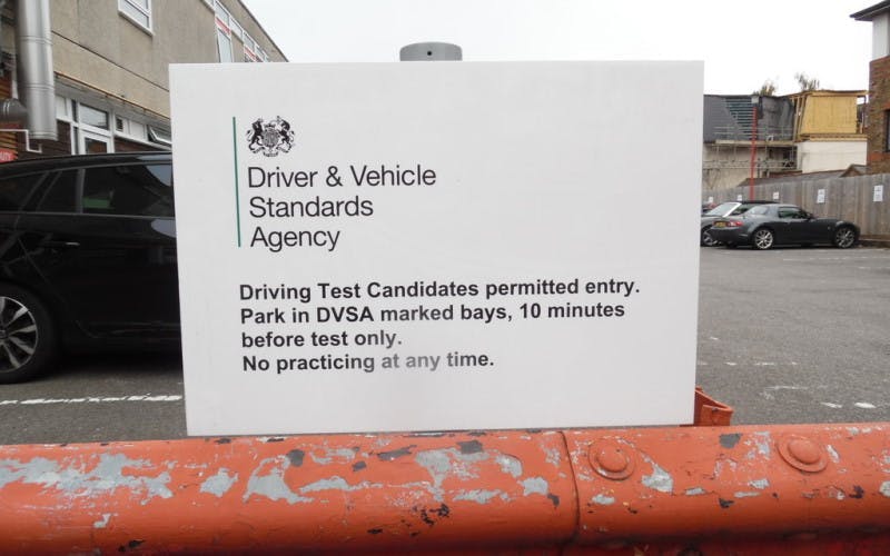 Photograph of a sign outside a DVSA test centre. The sign has the DVSA (Driver & Vehicle Standards Agency) logo in the top right. The body text of the sign says 'Driving Test Candidates permitted entry. Park in DVSA marked bays, 10 minutes before test only. No practicing at any time'