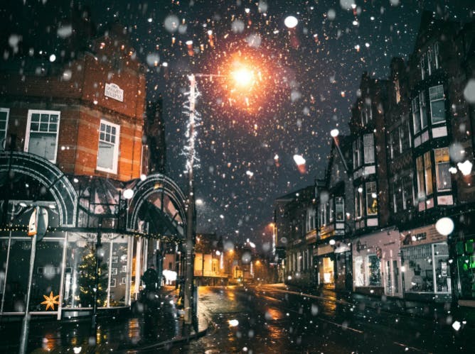 A UK street at night. It is snowing and Christmas decorations can be seen in windows
